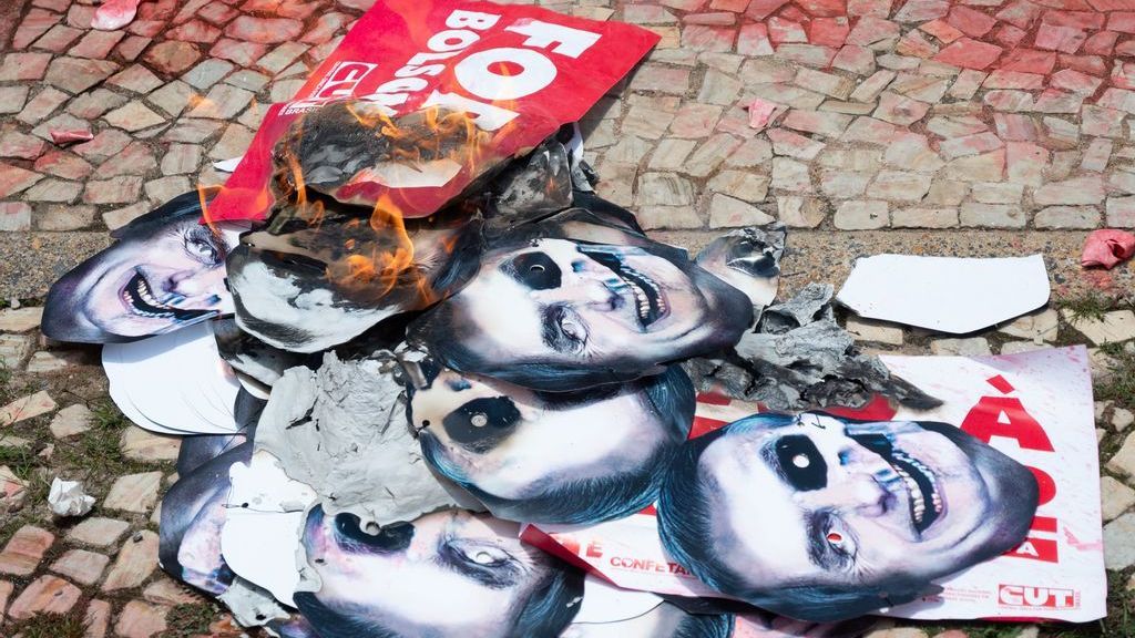 Masks with the altered image of Brazilian President Jair Bosonaro are burned during a protest about government measures to handle the COVID-19 pandemic on Oct. 20, in Brasilia, Brazil. The country’s Senate is currently looking into the government’s role in mismanaging the response to the virus. (Photo by Andressa Anholete/Getty Images)
